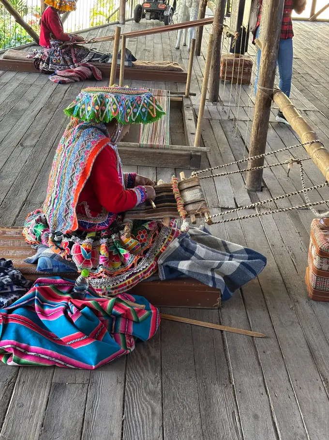 Frau in bunter Tracht und besticktem Hut sitzt auf einem Holzboden und webt an einem Webstuhl