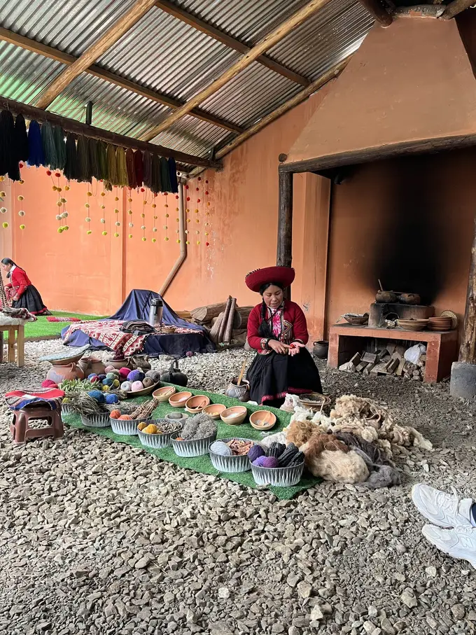 Eine Frau in traditioneller Tracht mit rotem Hut sitzt unter einem Wellblechdach vor Schalen mit gefärbter Wolle, Pflanzen und Rohwolle