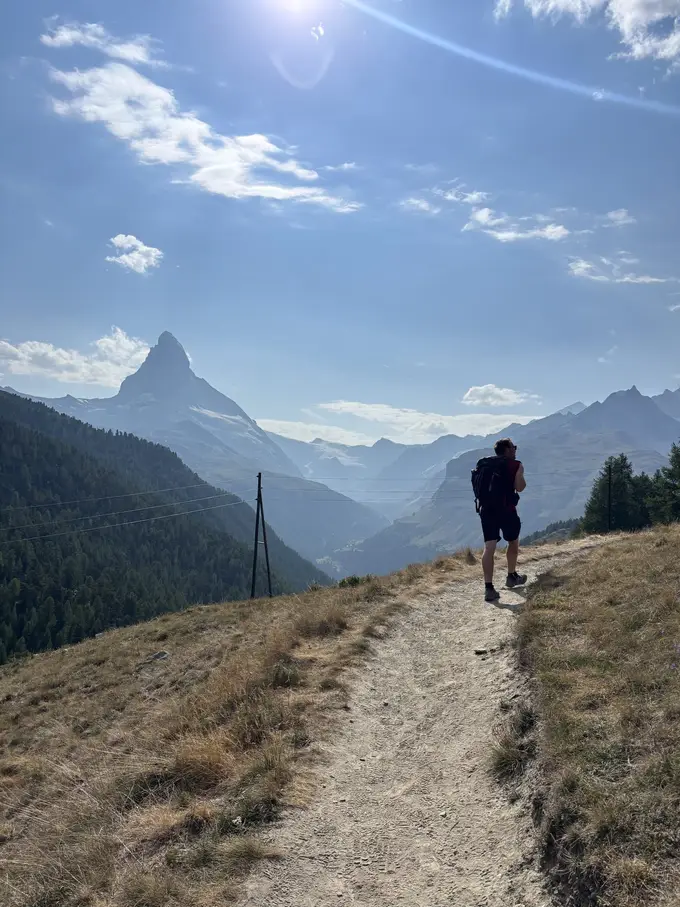Ein Wanderer mit Rucksack auf einem trockenen Pfad bergab, in der Ferne das Matterhorn im Gegenlicht