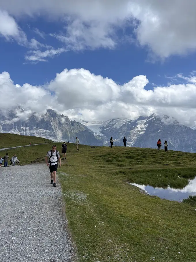 Max geht mit Rucksack auf einem breiten Schotterweg über eine Alpwiese, dahinter andere Wanderer und verschneite Berge in den Wolken