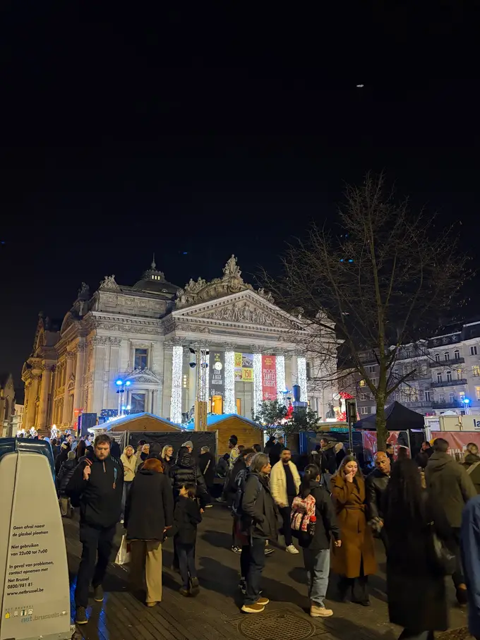 Die beleuchtete Börse bei Nacht mit lichterumwickelten Säulen, davor Holzbuden des Weihnachtsmarkts und viele Menschen