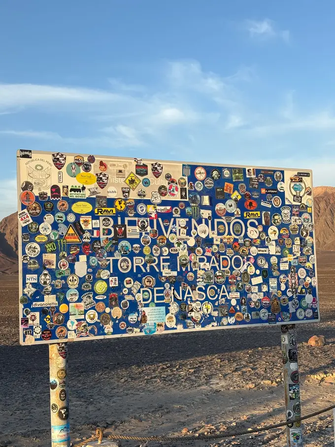 Blaues Schild „Bienvenidos Torre Mirador de Nasca“, fast vollständig mit Aufklebern aus aller Welt beklebt