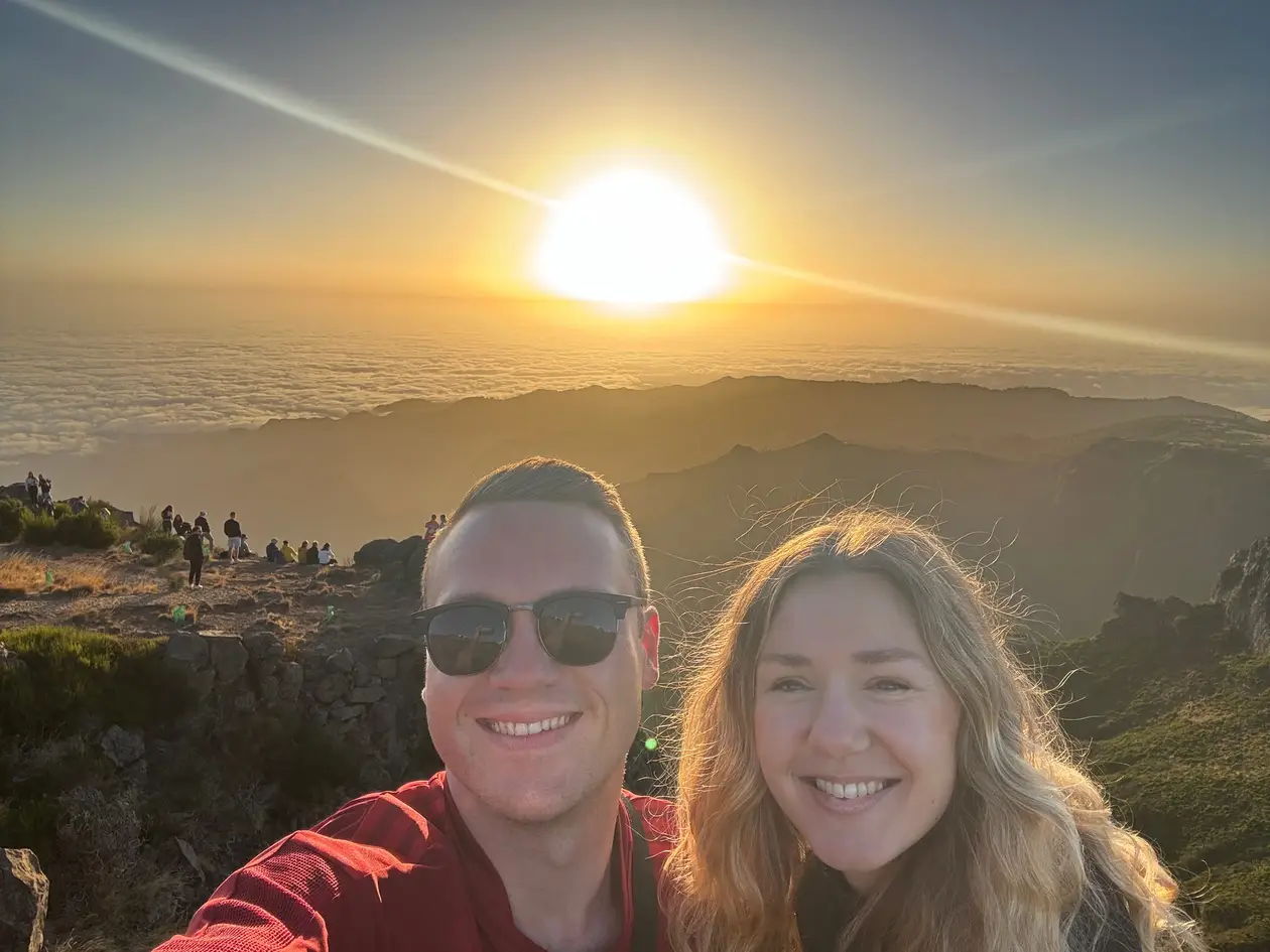 Max und Rebecca lächeln in die Kamera, dahinter geht am Pico do Arieiro die Sonne über einem geschlossenen Wolkenmeer auf