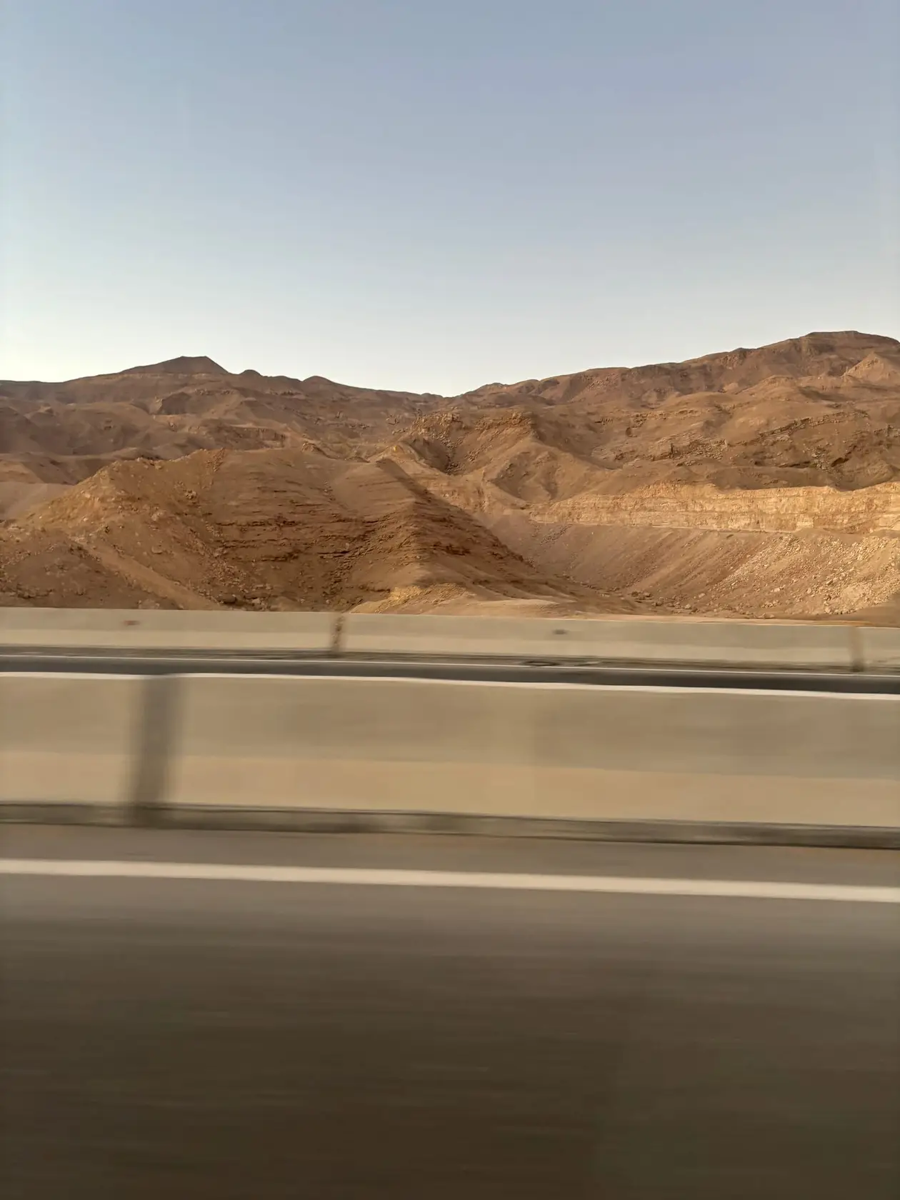 Verwischter Blick aus dem fahrenden Bus auf eine Schnellstraße mit Betonleitplanke, dahinter kahle, braune Wüstenberge