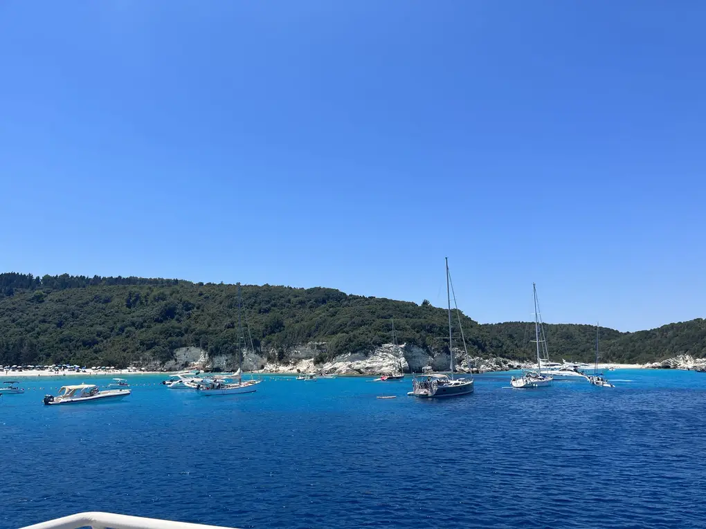 Motorboote und Segelyachten ankern vor einem hellen Strand unter einem bewaldeten Hügel
