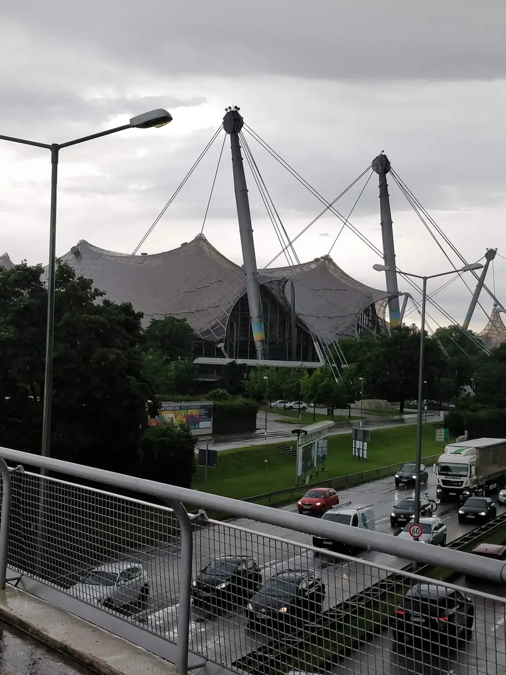 Das geschwungene Zeltdach im Olympiapark mit hohen Masten und Stahlseilen, davor eine mehrspurige Straße mit Autos