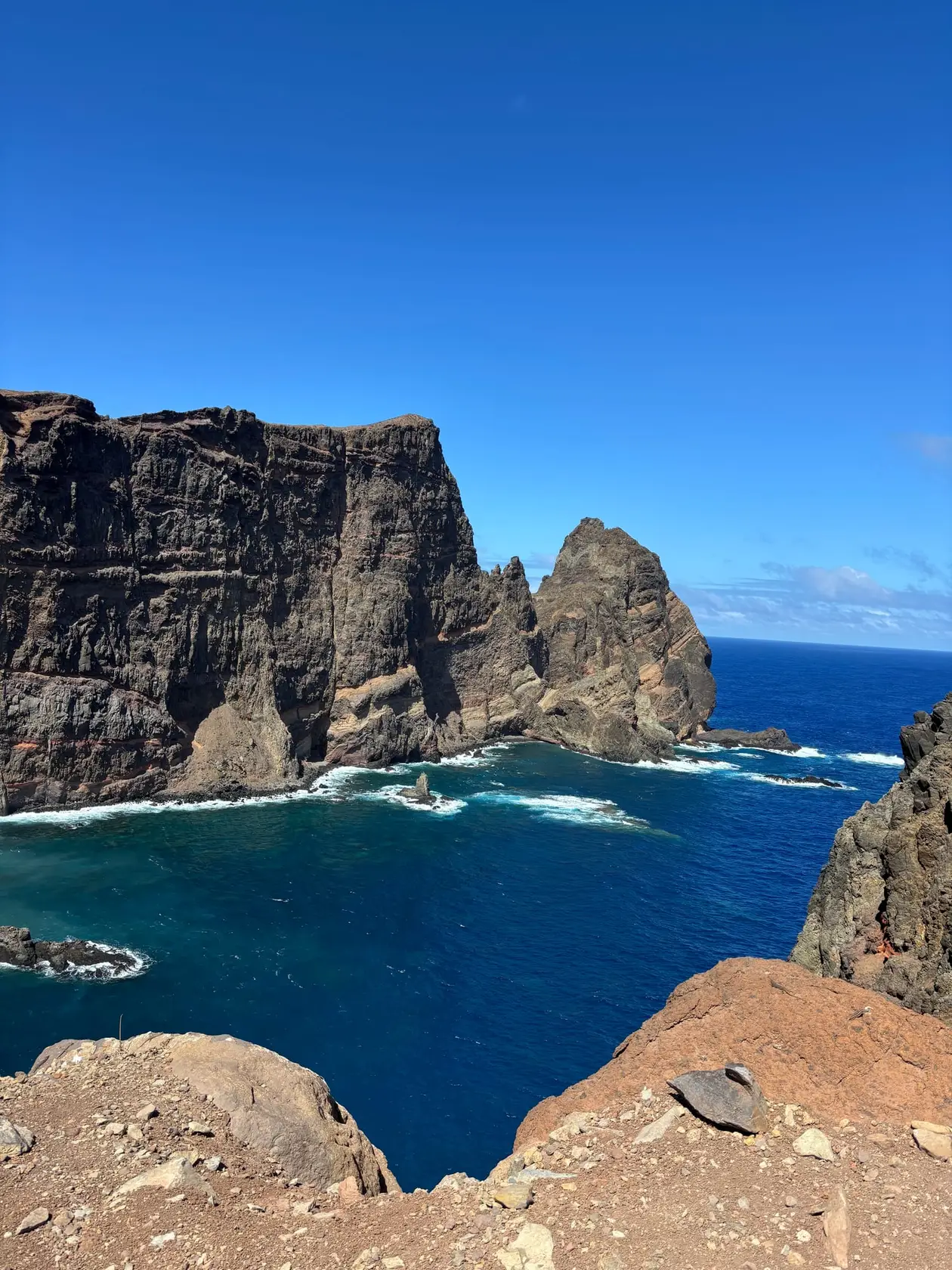 Steile rot-braune Felsklippen ragen aus dem tiefblauen Atlantik, Wellen brechen am Fuß der Klippen