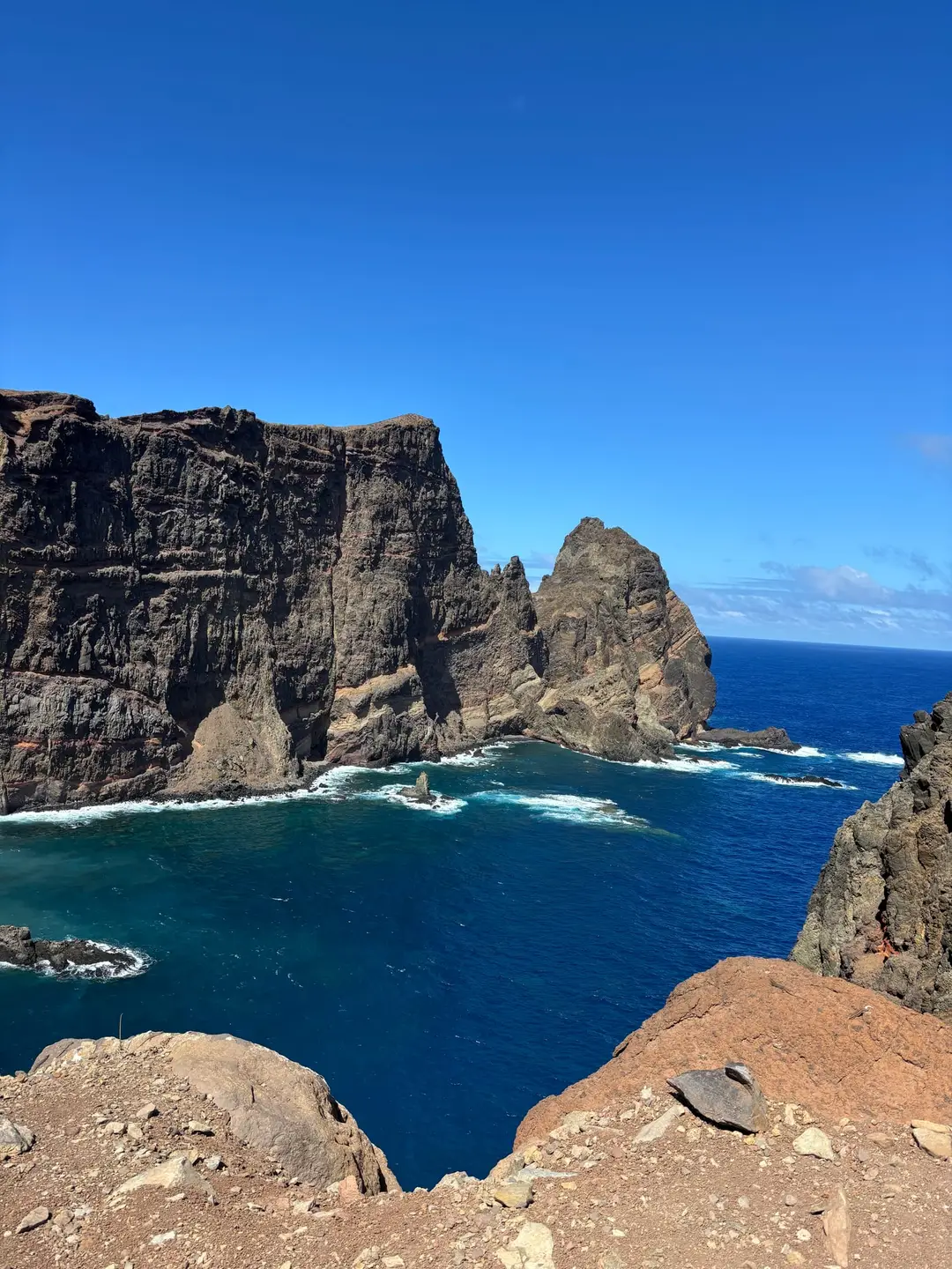 Steile rot-braune Felsklippen ragen aus dem tiefblauen Atlantik, Wellen brechen am Fuß der Klippen