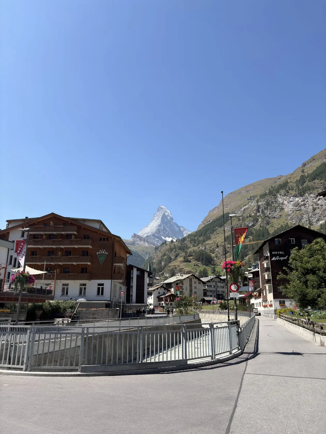 Eine Straße mit Holzhäusern und Hotels in Zermatt, am Ende das Matterhorn unter blauem Himmel