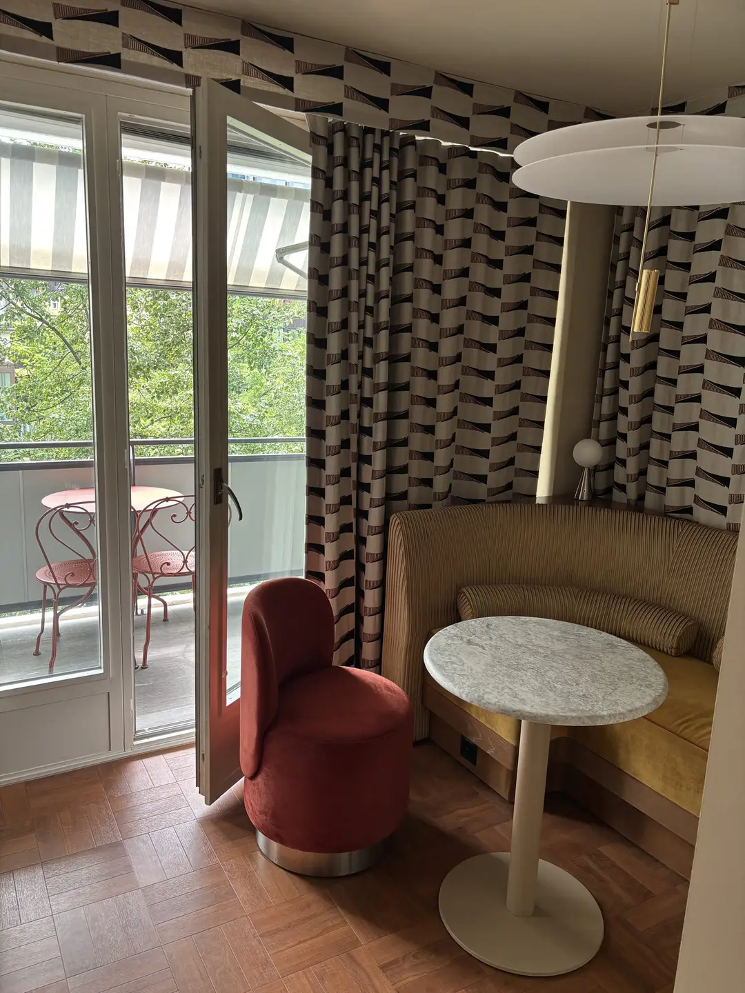 Hotelzimmer mit rotem Sessel, rundem Marmortisch und gepolsterter Sitzbank, dahinter eine offene Glastür zum Balkon mit zwei roten Metallstühlen und Markise