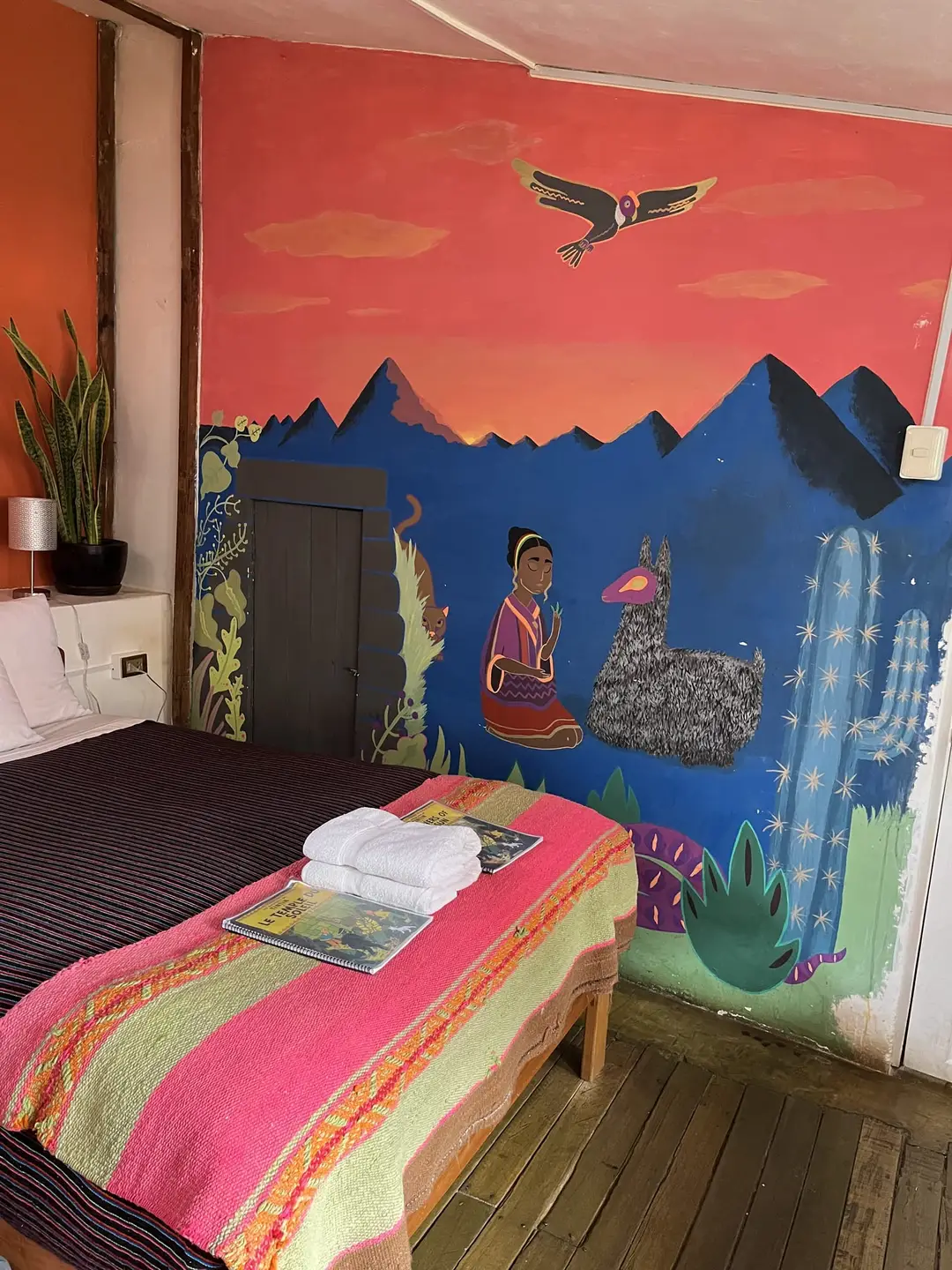 Doppelbett mit pinker Wolldecke vor einem Wandbild mit Bergen, einem Kondor, Kakteen und einer Frau mit Lama