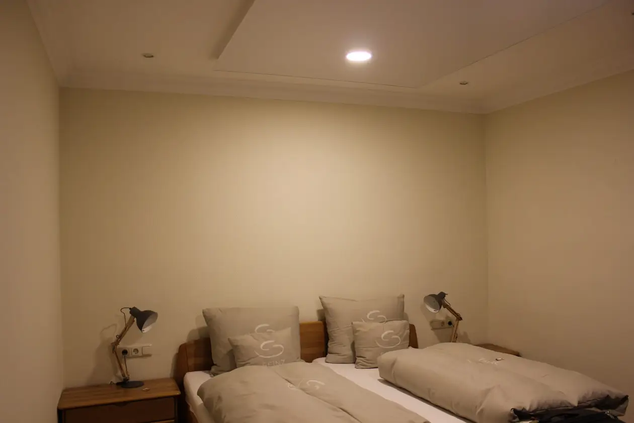 Hotelzimmer mit hellen Wänden, einem Doppelbett aus Holz mit beigefarbener Bettwäsche und zwei Schreibtischlampen auf den Nachttischen