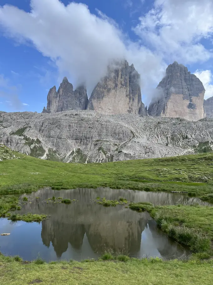 Die Drei Zinnen mit Wolken an den Spitzen spiegeln sich in einem kleinen Tümpel auf einer Almwiese